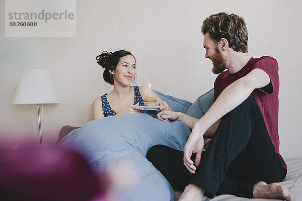 Glückliche junge Frau hält Geburtstagskuchen mit Kerze  während sie den Mann im Bett anschaut