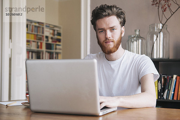 Porträt eines selbstbewussten jungen Mannes mit Laptop am Tisch