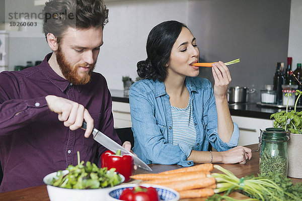 Junger Mann hackt rote Paprika neben einer Frau  die Karotte am Küchentisch isst.