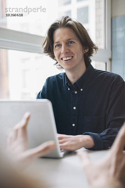 Junger Mann lächelt Kollegen an  während er den Laptop am Tisch benutzt.