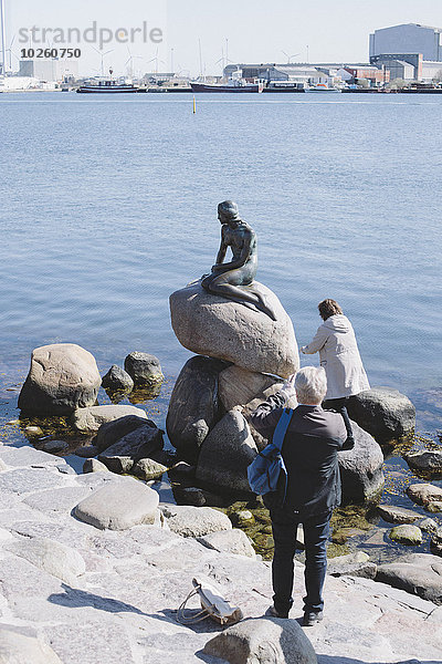 Menschen am Seeufer mit Statue auf Felsen