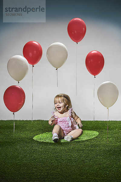 Fröhliches Mädchen sitzt auf Gras mit roten und weißen Luftballons in der Luft.