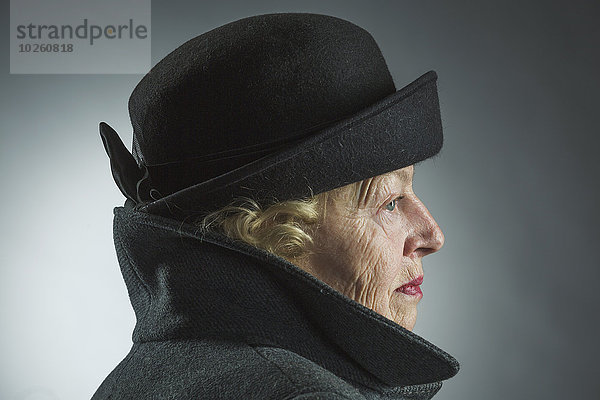 Seitenansicht der Seniorin mit Hut und Jacke vor grauem Hintergrund