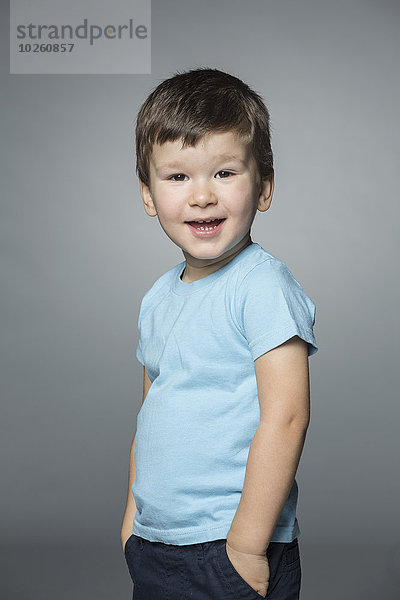 Porträt eines glücklichen Jungen mit Händen in den Taschen vor grauem Hintergrund