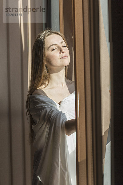 Junge Frau in Laken gehüllt  genießt Sonnenlicht durchs Fenster
