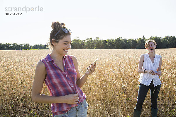 Zwei junge Frauen stehen in einem Maisfeld  eine hält ihr Handy in der Hand.