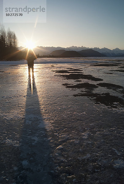 Junger Mann auf einem zugefrorenen See stehend  mit erhobenen Armen und Sonnenstrahlen über dem Kopf; Haines  Alaska  Vereinigte Staaten von Amerika'.