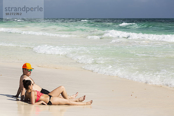 Paar liegt und sitzt am Sandstrand mit einlaufenden Wellen und blauem Himmel; Tulum  Quintana Roo  Mexiko'.