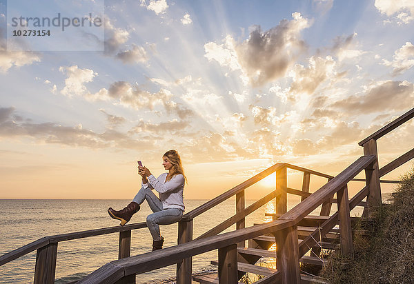 Eine Frau sitzt auf einem Holzgeländer am Wasser und telefoniert mit einem Handy; Tarifa  Cadiz  Andalusien  Spanien'.