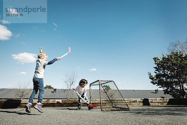 Aufgeregtes Mädchen spielt Hockey mit dem Jungen auf dem Hof gegen den blauen Himmel.