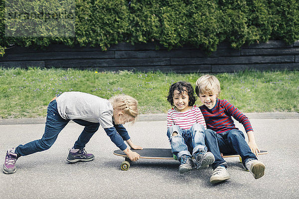 Mädchen schiebt Freunde  die auf dem Skateboard auf dem Hof sitzen.