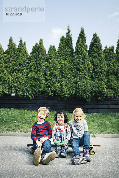 Porträt von glücklichen Kindern  die auf einem Skateboard gegen Bäume sitzen.