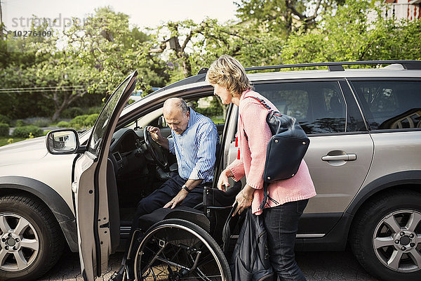 Behinderter Mann beim Aussteigen aus dem Auto auf einem Rollstuhl  der von einer Frau gehalten wird.