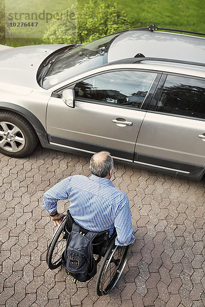 Hochwinkelansicht eines reifen Mannes im Rollstuhl  der sich auf der Straße in Richtung Auto bewegt.