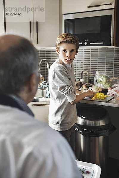 Junge schaut Vater an  während er in der Küche Gemüse schneidet.