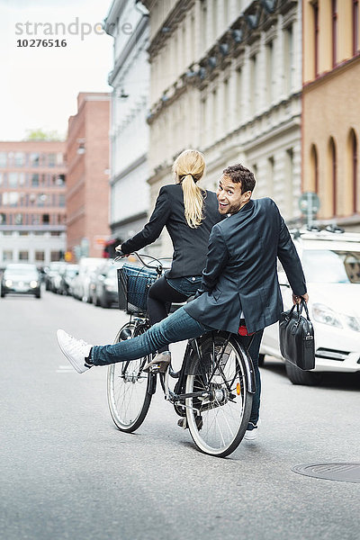 Rückansicht Porträt eines glücklichen Geschäftsmannes  der auf dem Rücksitz sitzt  während eine Kollegin Fahrrad fährt.