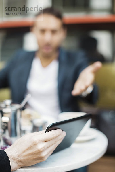 Geschäftsmann gestikuliert gegenüber einer Frau  die ein digitales Tablett im Straßencafé hält.