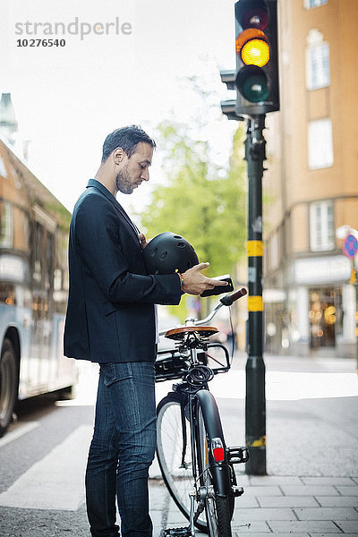 Seitenansicht des Geschäftsmannes mit dem Handy im Stehen mit dem Fahrrad auf der Stadtstraße