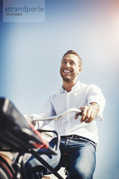 Flachwinkelansicht eines glücklichen Geschäftsmannes auf dem Fahrrad gegen den blauen Himmel