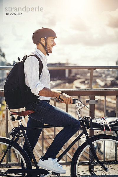 Lächelnder Geschäftsmann mit Fahrrad und Blick auf die Stadt  während er auf der Brücke steht.
