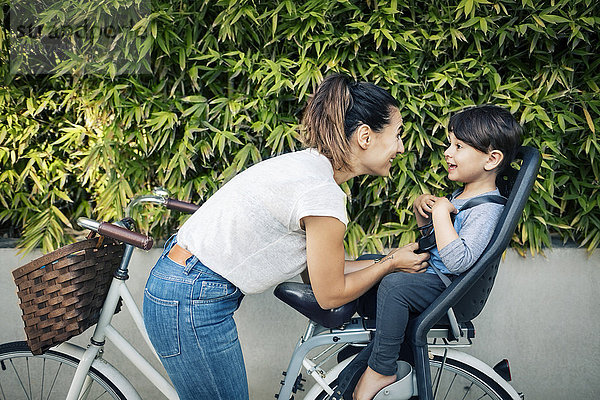 Glückliche Frau schaut auf den Sohn  der auf dem Rücksitz des Fahrrads gegen die Pflanzen sitzt.