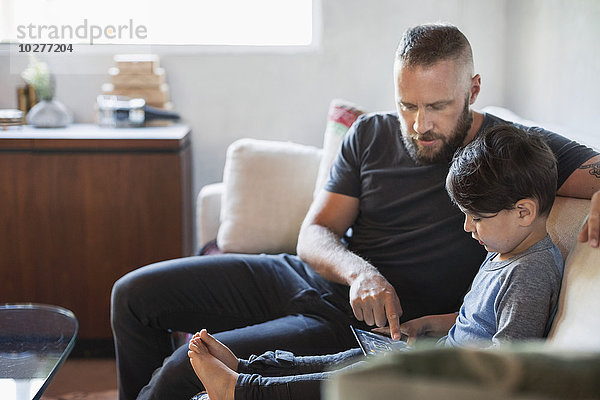 Vater hilft dem Sohn bei der Verwendung des digitalen Tabletts auf dem Sofa zu Hause.