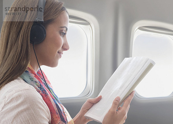 Junge Frau trägt Kopfhörer beim Lesen eines Buches im Flugzeug