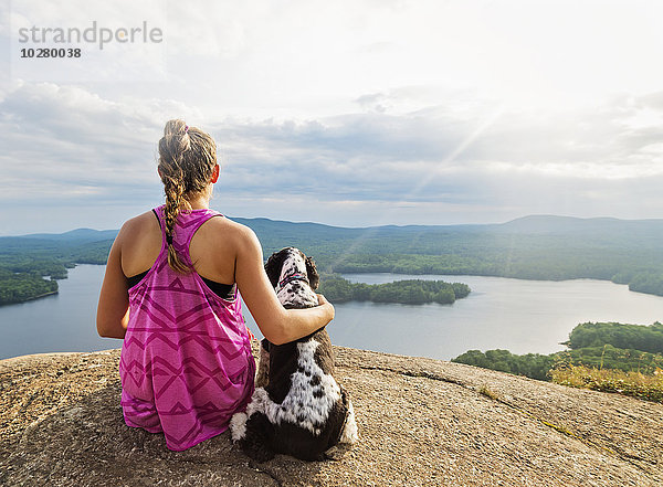 Junge Frau sitzt mit Hund auf einer Klippe mit Blick auf den See