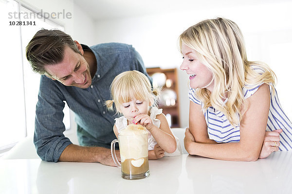 Mädchen (2-3) trinkt Milchshake mit Eltern zu Hause