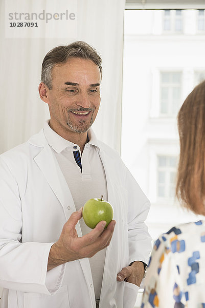 Männlicher Arzt bietet einem Patienten in seiner Klinik einen Apfel an  München  Bayern  Deutschland