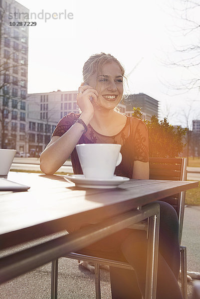 Junge Frau spricht in einem Straßencafé mit einem Mobiltelefon  München  Bayern  Deutschland