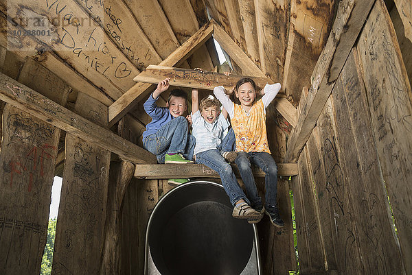 Drei Kinder sitzen auf dem Holzbalken eines Baumhauses auf einem Spielplatz  München  Bayern  Deutschland