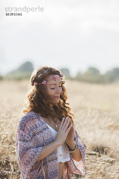 Serene boho Frau meditiert mit den Händen in der Mitte des Herzens in einem sonnigen ländlichen Gebiet.