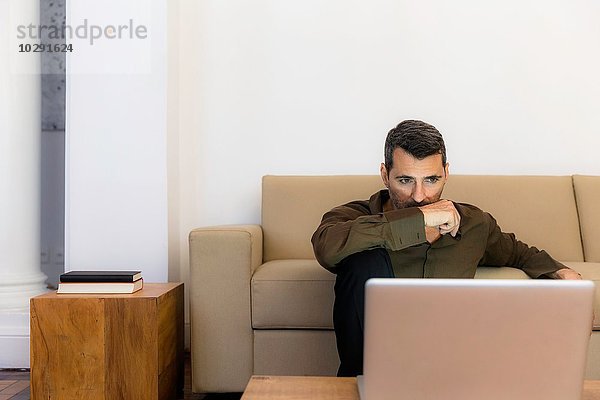 Älterer Mann auf dem Boden sitzend mit Blick auf Laptop  Arm über Mund