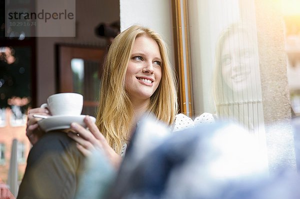 Junge Frau mit langen blonden Haaren sitzend mit Kaffeetasse  lächelnd wegblickend