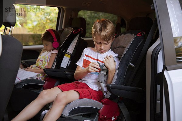 Mädchen mit digitalem Tablett  während der Bruder den Sicherheitsgurt auf dem Rücksitz des Autos anschnallt.