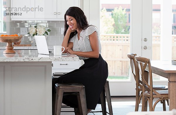 Junge Frau  sitzend an der Küchentheke mit Kaffee und Laptop