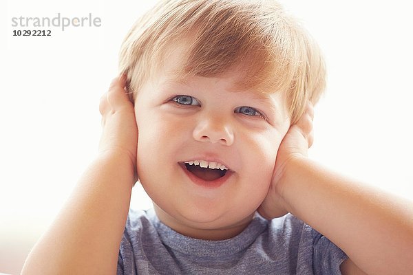 Porträt eines kleinen Jungen  der die Ohren mit den Händen bedeckt und lächelnd wegsieht.