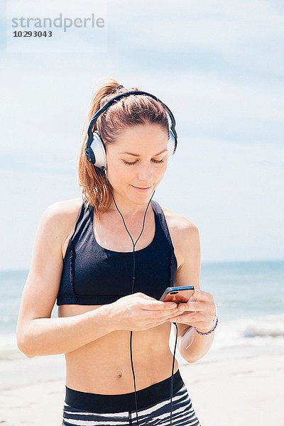 Mittlere erwachsene Frau  die am Strand steht  Kopfhörer trägt und einen mp3-Player ansieht.