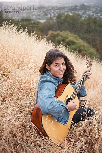 Porträt einer jungen Frau  die auf einem grasbewachsenen Hügel Akustikgitarre spielt.