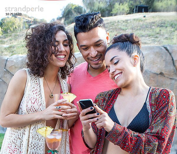 Junger Mann und zwei Schwestern beim Lesen von Smartphone-Texten im Garten