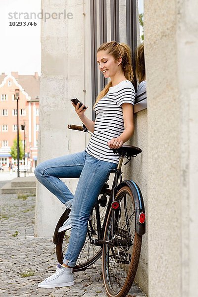 Seitenansicht der jungen Frau  die das Fahrrad an der Gebäudeaußenseite anlehnt und das Smartphone lächelnd betrachtet.