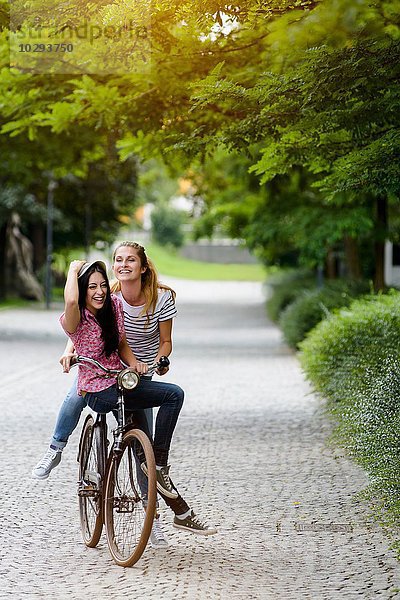 Vorderansicht von jungen Frauen  die sich eine Fahrradtour teilen und sich am Hut festhalten.
