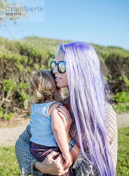 Junge Frau mit langen violetten Haaren  die eine kleine Tochter hält.