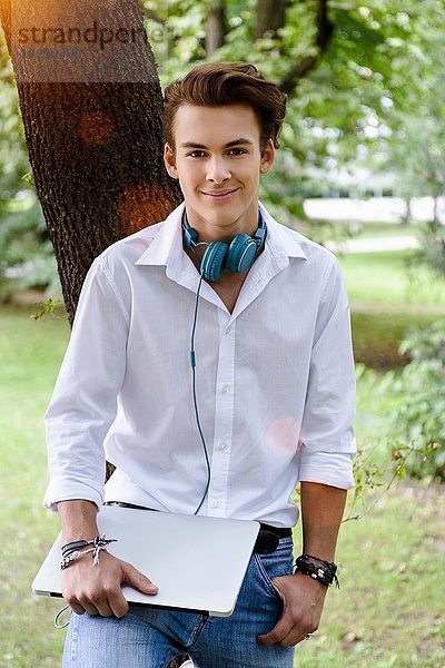 Junger Mann mit Kopfhörer an Baum gelehnt  Hand in Tasche  hält Laptop mit Blick auf die Kamera lächelnd