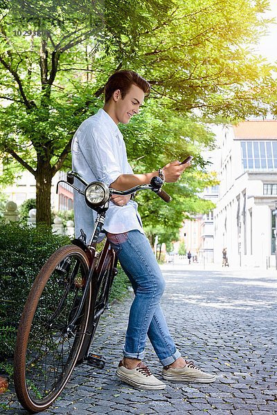 Seitenansicht eines jungen Mannes  der sich gegen das Fahrrad lehnt und das Smartphone lächelnd ansieht.