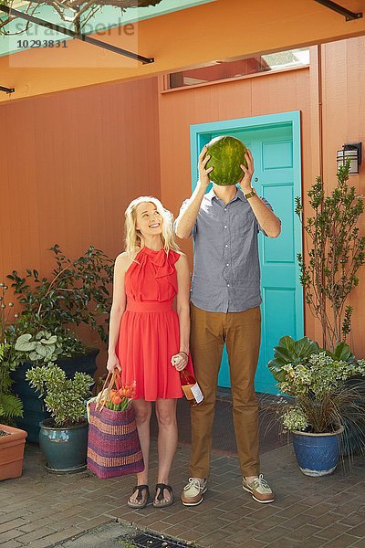 Frau mit Weinflasche und Mann  der das Gesicht mit Wassermelone bedeckt.