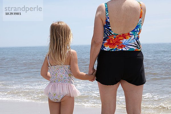 Rückansicht der älteren Frau und Enkelin  die am Strand Händchen halten.