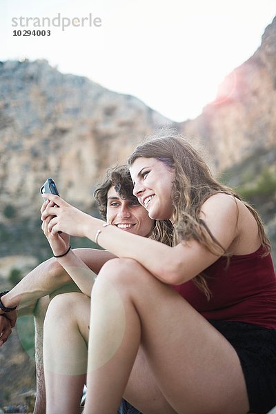 Junger Mann und jugendliche Schwester beim Lesen von Smartphone-Texten am felsigen Strand  Javea  Spanien