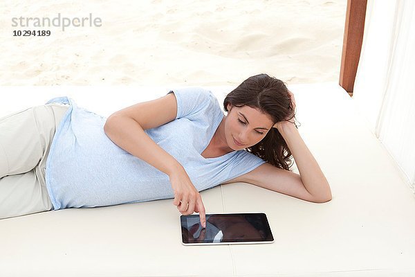 Schwangere mittlere erwachsene Frau  die auf einer Liege am Strand liegt und ein digitales Tablett benutzt.